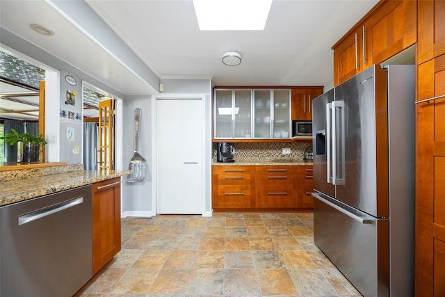 kitchen featuring stainless steel appliances, glass insert cabinets, backsplash, brown cabinetry, and light stone countertops