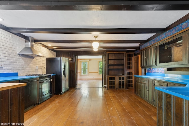 kitchen featuring black gas range, wall chimney range hood, stainless steel fridge, light wood finished floors, and beamed ceiling
