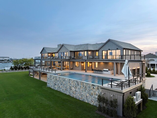 34 Westbridge Rd, Westhampton Beach, NY 11978 - back of house at dusk featuring a balcony, a lawn, an outdoor pool, an outdoor hangout area, and a patio
