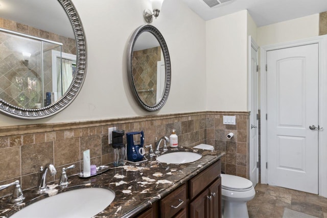full bath featuring tile walls, a stall shower, toilet, double vanity, and a wainscoted wall