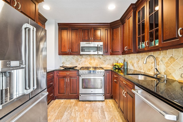kitchen with stainless steel appliances, a sink, dark stone counters, decorative backsplash, and glass insert cabinets