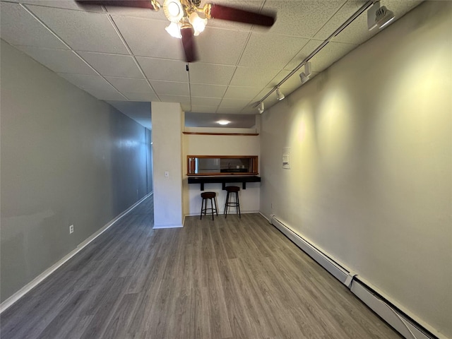 below grade area with baseboard heating, wood finished floors, a ceiling fan, a drop ceiling, and baseboards