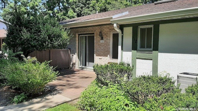 entrance to property featuring fence, brick siding, roof with shingles, cooling unit, and covered porch