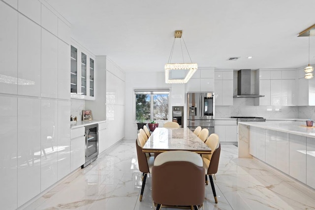 kitchen with modern cabinets, stainless steel fridge with ice dispenser, wall chimney range hood, beverage cooler, and light marble finish floors