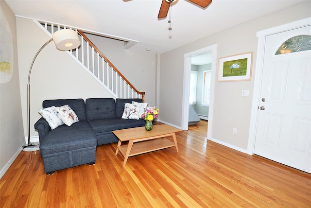 living room with light wood-type flooring, ceiling fan, stairs, baseboard heating, and baseboards
