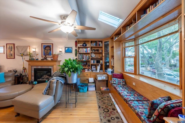 living area with light wood finished floors, ceiling fan, a tiled fireplace, and a skylight