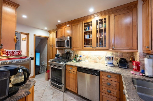 kitchen with appliances with stainless steel finishes, brown cabinetry, light tile patterned floors, backsplash, and light stone counters