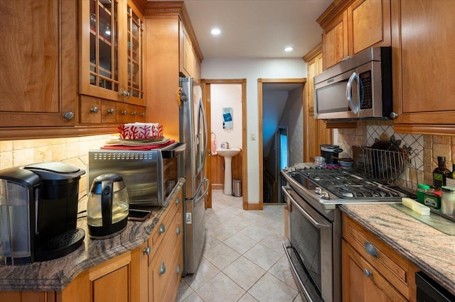 kitchen featuring appliances with stainless steel finishes, brown cabinets, decorative backsplash, dark stone counters, and glass insert cabinets