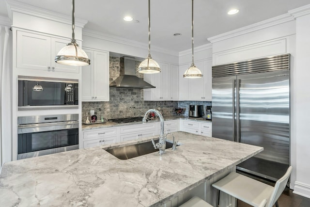 kitchen featuring built in appliances, wall chimney exhaust hood, a sink, backsplash, and white cabinets