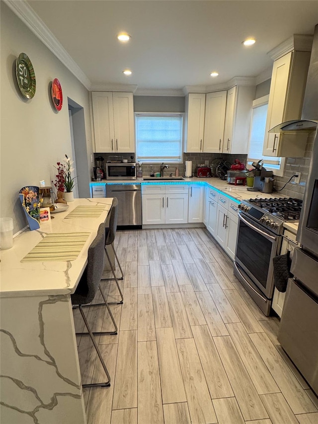 kitchen with appliances with stainless steel finishes, wall chimney range hood, a sink, ornamental molding, and white cabinetry
