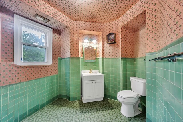 bathroom with tile walls, wallpapered walls, a wainscoted wall, toilet, and vanity