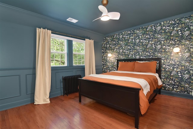 bedroom featuring radiator heating unit, wallpapered walls, wood finished floors, ornamental molding, and a wainscoted wall