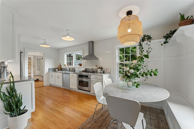 kitchen featuring wall chimney exhaust hood, appliances with stainless steel finishes, white cabinets, light wood-style flooring, and tasteful backsplash