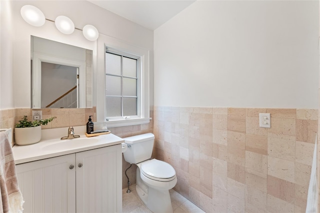 half bath with toilet, tile walls, wainscoting, vanity, and tile patterned flooring