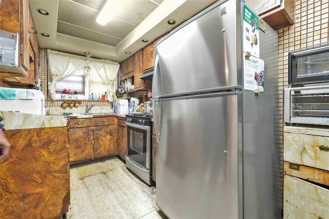 kitchen with stainless steel appliances, under cabinet range hood, brown cabinetry, a tray ceiling, and light countertops