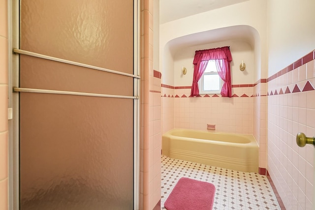 bathroom featuring a shower with shower door, tile walls, a wainscoted wall, and a tub