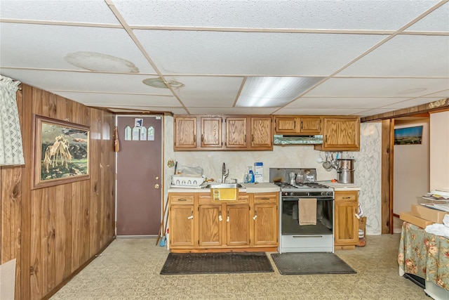 kitchen with gas range oven, under cabinet range hood, a sink, wood walls, and light countertops