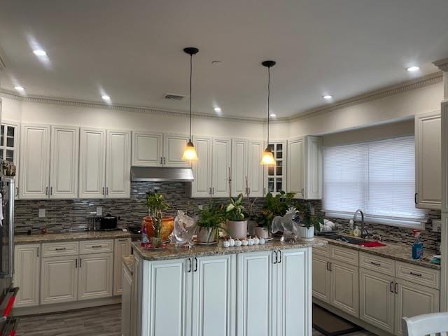 kitchen featuring under cabinet range hood, a sink, decorative backsplash, a kitchen island, and ornamental molding