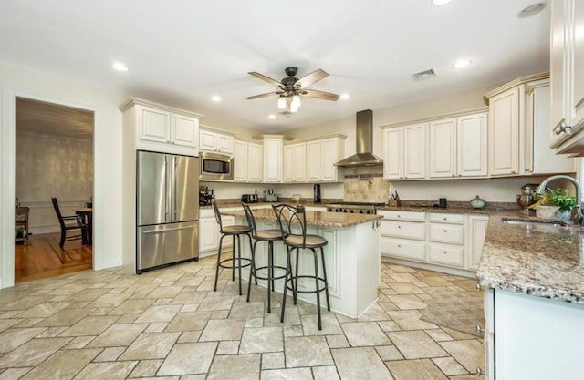 kitchen with appliances with stainless steel finishes, wall chimney exhaust hood, a sink, ceiling fan, and a breakfast bar