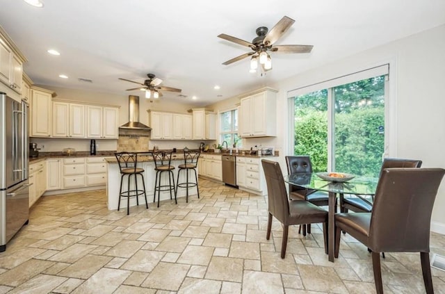 kitchen featuring wall chimney exhaust hood, stainless steel appliances, a ceiling fan, cream cabinets, and a kitchen bar