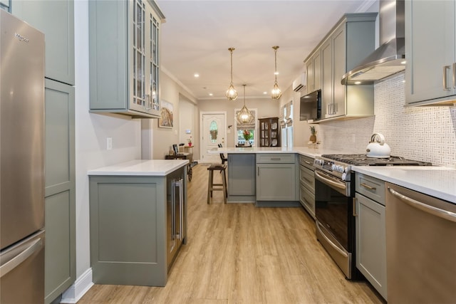 kitchen featuring appliances with stainless steel finishes, wall chimney exhaust hood, a peninsula, light wood finished floors, and a kitchen bar