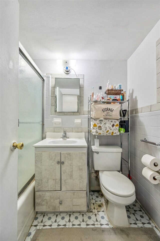 bathroom with tile walls, tile patterned floors, toilet, shower / bath combination with glass door, and vanity