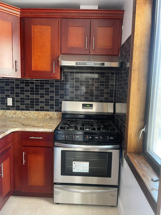 kitchen with stainless steel gas range, under cabinet range hood, backsplash, light stone countertops, and light tile patterned floors