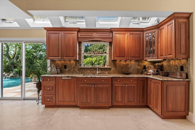 kitchen with a skylight, a sink, light stone counters, backsplash, and brown cabinets