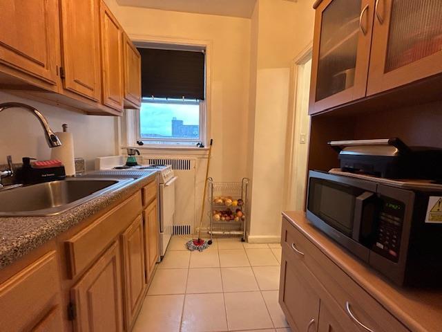 86-10 34th Ave Unit 624, Jackson Heights, NY 11372 - kitchen featuring stainless steel microwave, a sink, light tile patterned floors, brown cabinetry, and glass insert cabinets
