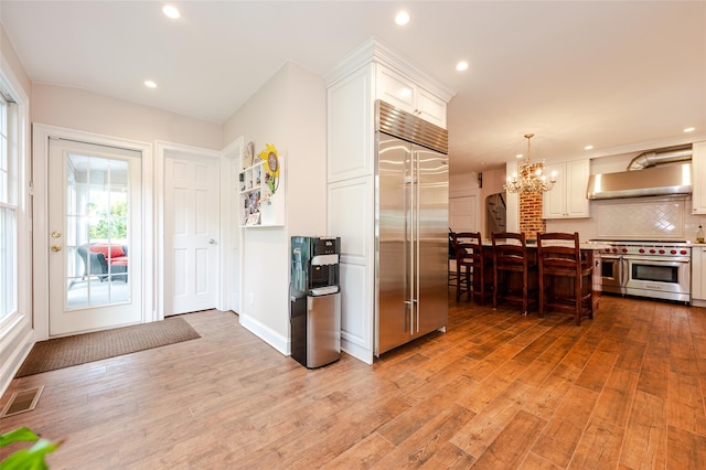 70 Loines Ave, Merrick, NY 11566 - kitchen with premium appliances, wall chimney exhaust hood, a chandelier, decorative backsplash, and light wood-type flooring
