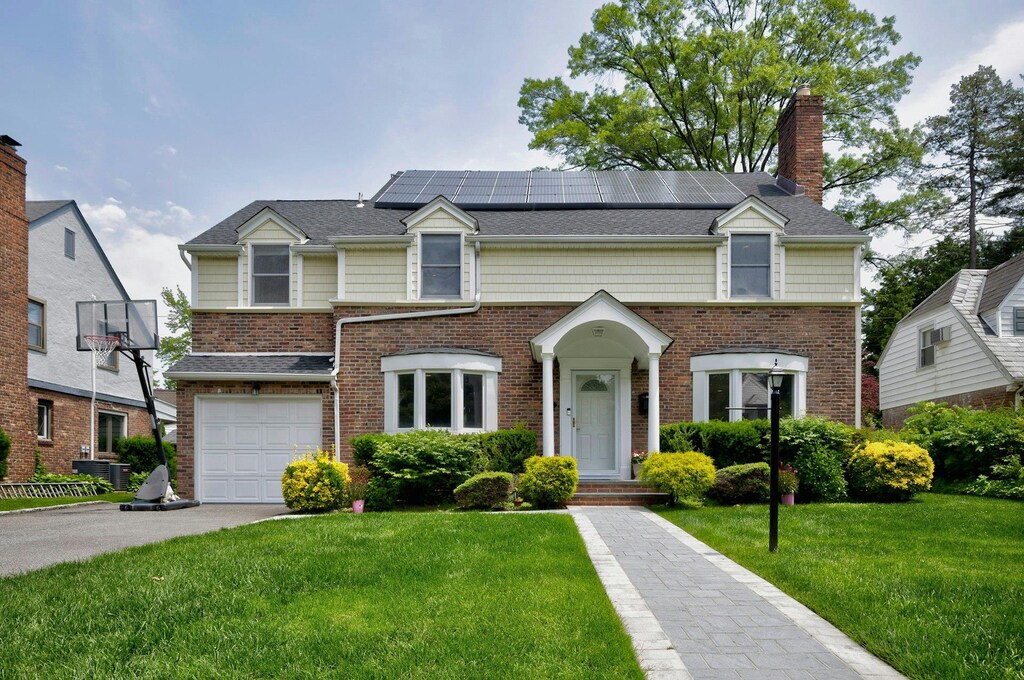 16 Andover Ln, Manhasset, NY 11030 - view of property featuring a front yard, a chimney, roof mounted solar panels, driveway, and a garage