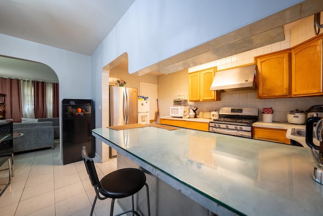 32-23 76th St, East Elmhurst, NY 11370 - kitchen with premium range hood, appliances with stainless steel finishes, a kitchen breakfast bar, arched walkways, and light tile patterned floors