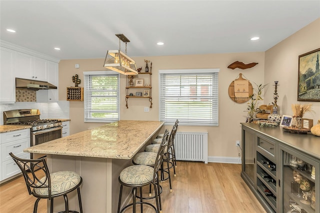 249-14 57th Ave Unit 259, Little Neck, NY 11362 - kitchen featuring stainless steel gas stove, under cabinet range hood, radiator heating unit, a kitchen breakfast bar, and white cabinetry