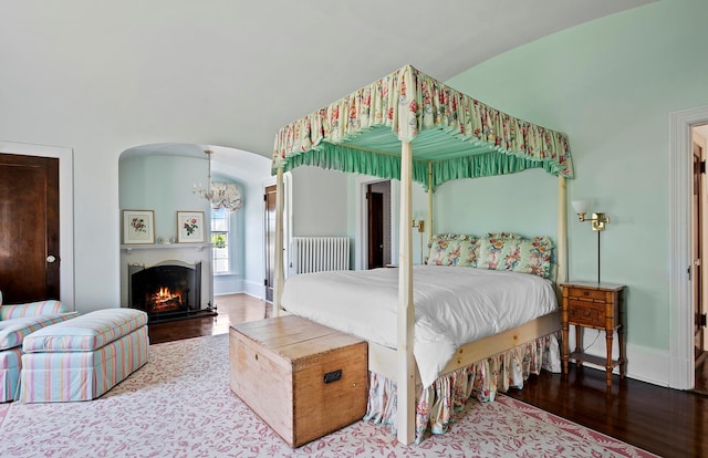 107 Quogue St, Quogue, NY 11959 - bedroom with wood finished floors, a chandelier, arched walkways, radiator heating unit, and a warm lit fireplace