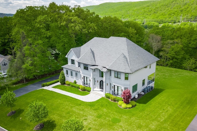 view of front facade featuring a view of trees, a front lawn, driveway, a shingled roof, and stucco siding