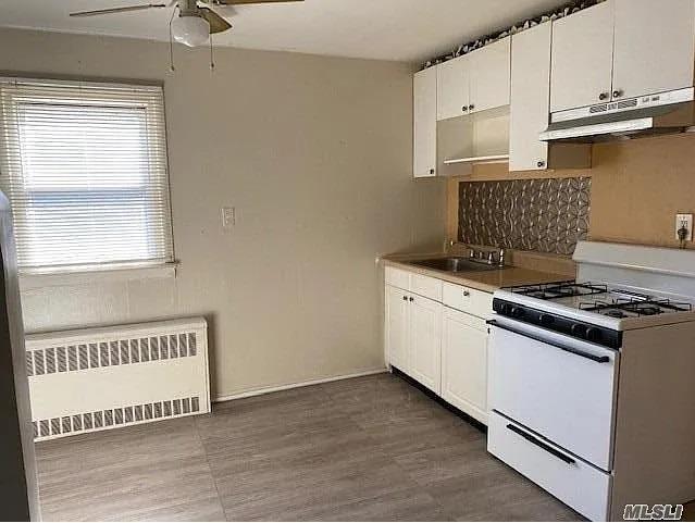 116-46 171st St, Jamaica, NY 11434 - kitchen featuring white gas range, under cabinet range hood, radiator, a ceiling fan, and white cabinetry