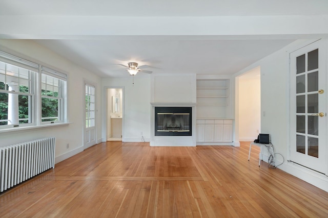 1459 N Country Rd, Wading River, NY 11792 - unfurnished living room featuring radiator, light wood finished floors, ceiling fan, built in shelves, and a glass covered fireplace