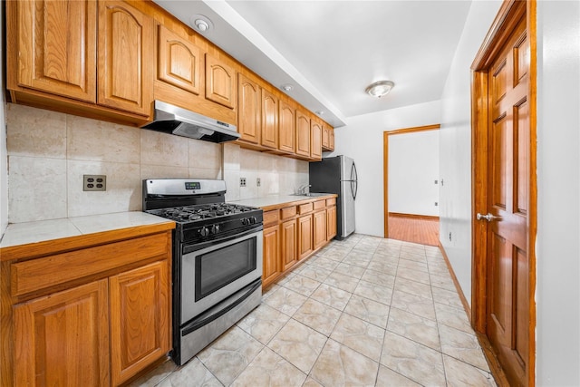 2162 24th St, Astoria, NY 11105 - kitchen featuring appliances with stainless steel finishes, under cabinet range hood, backsplash, tile countertops, and brown cabinets