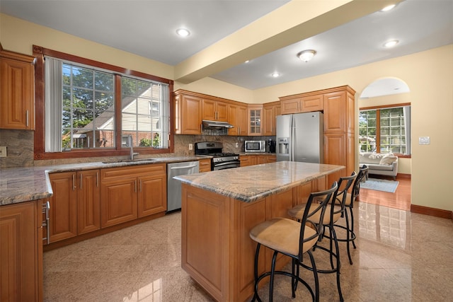 83-14 266th St, Glen Oaks, NY 11004 - kitchen featuring appliances with stainless steel finishes, under cabinet range hood, stone counters, brown cabinetry, and a kitchen breakfast bar