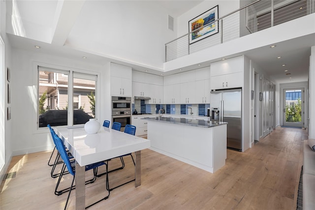 846 Dune Rd, Westhampton Dunes, NY 11978 - kitchen with stainless steel appliances, a towering ceiling, white cabinets, light wood-style floors, and a kitchen island with sink
