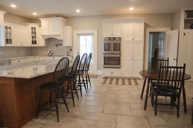 kitchen featuring a kitchen breakfast bar, double oven, light stone countertops, decorative backsplash, and white cabinets