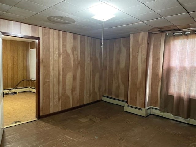 empty room featuring a drop ceiling, wooden walls, and a baseboard heating unit