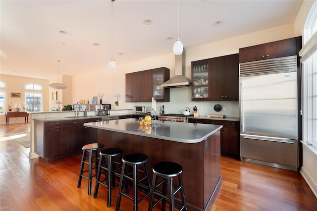 kitchen featuring a breakfast bar area, stainless steel appliances, dark brown cabinets, wall chimney exhaust hood, and glass insert cabinets