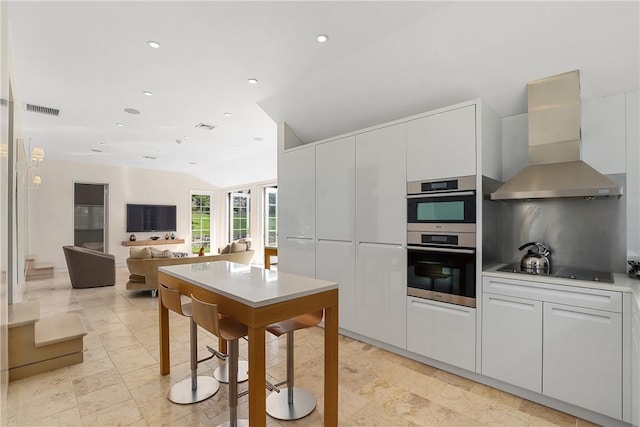 kitchen featuring wall chimney exhaust hood, black electric cooktop, double oven, white cabinets, and lofted ceiling