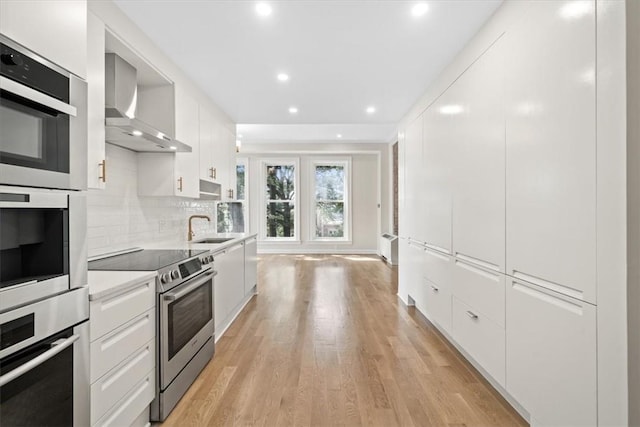 kitchen with appliances with stainless steel finishes, white cabinetry, sink, light hardwood / wood-style floors, and wall chimney range hood