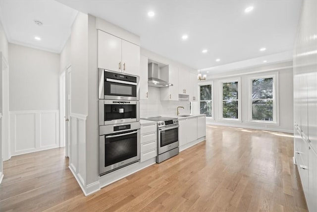 kitchen with wall chimney range hood, light hardwood / wood-style flooring, appliances with stainless steel finishes, white cabinetry, and decorative backsplash