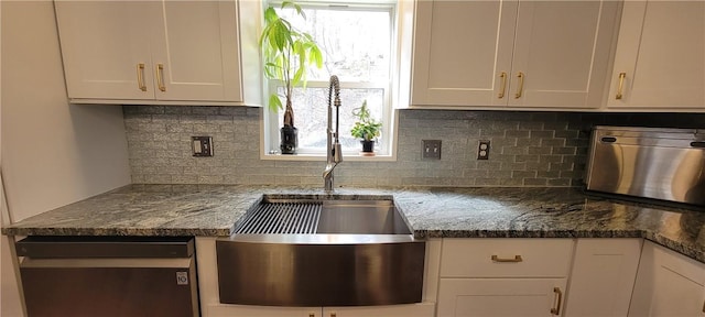 kitchen with a sink, dark stone countertops, backsplash, stainless steel dishwasher, and white cabinets