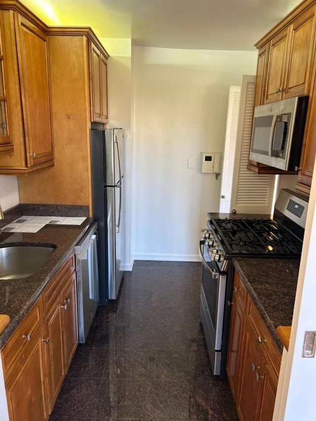 kitchen with baseboards, brown cabinets, a sink, dark stone countertops, and appliances with stainless steel finishes