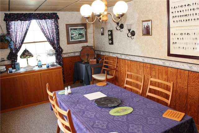 carpeted dining space with a paneled ceiling, wood walls, and an inviting chandelier