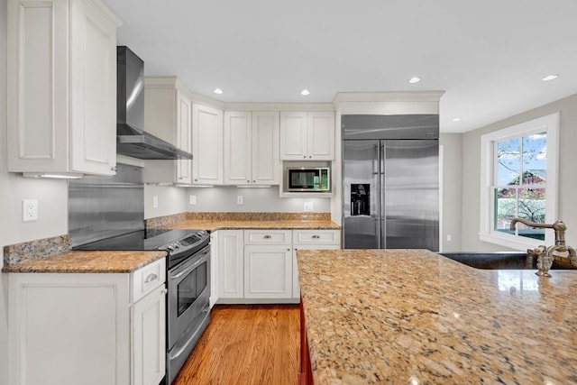 kitchen with white cabinetry, light stone countertops, wall chimney range hood, light hardwood / wood-style flooring, and built in appliances